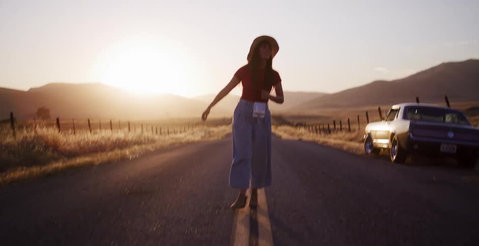 A stylish young woman playfully walks and dances down the middle of a deserted country road as the sun sets, with a vintage muscle car parked on the shoulder