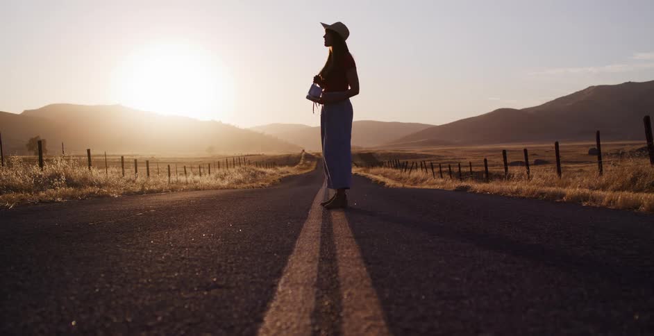 A silhouetted woman walks along the center line of a deserted country road at sunrise before stopping to take a picture of the beautiful landscape