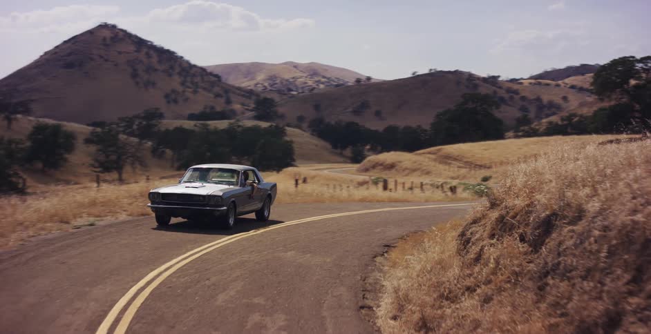 A classic Ford Mustang cruises along a winding country road through sun-drenched golden hills in slow motion