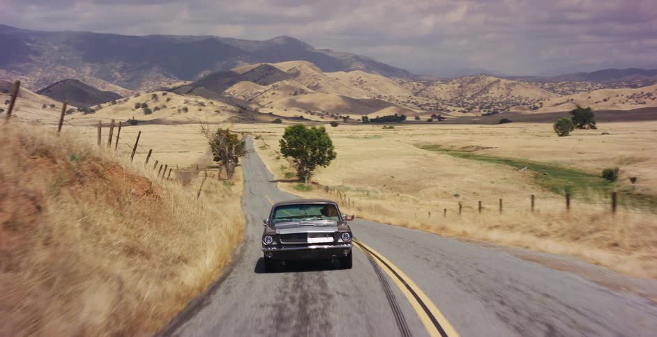 A classic gray muscle car drives along a winding country road through a vast, dry, hilly landscape
