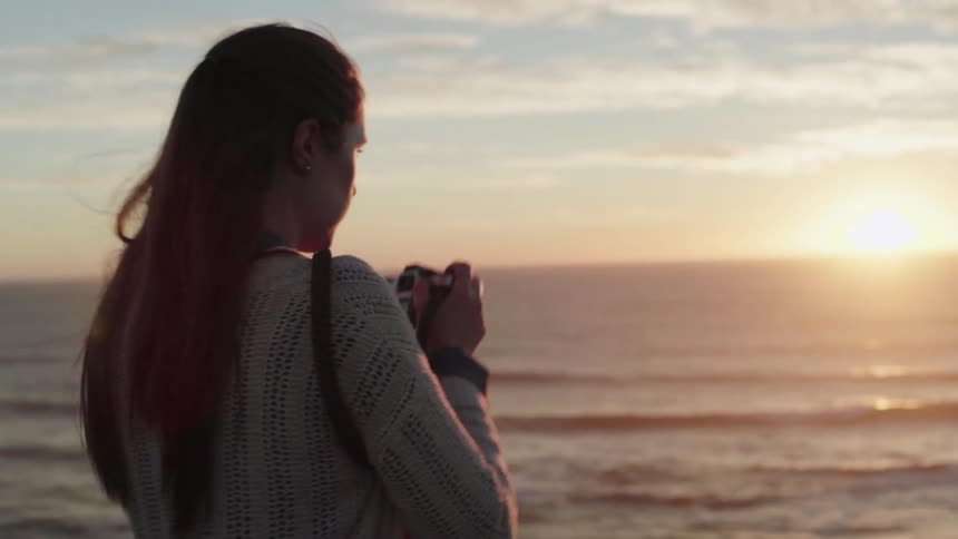 A woman takes a photo and lowers her camera as she stand at the beach