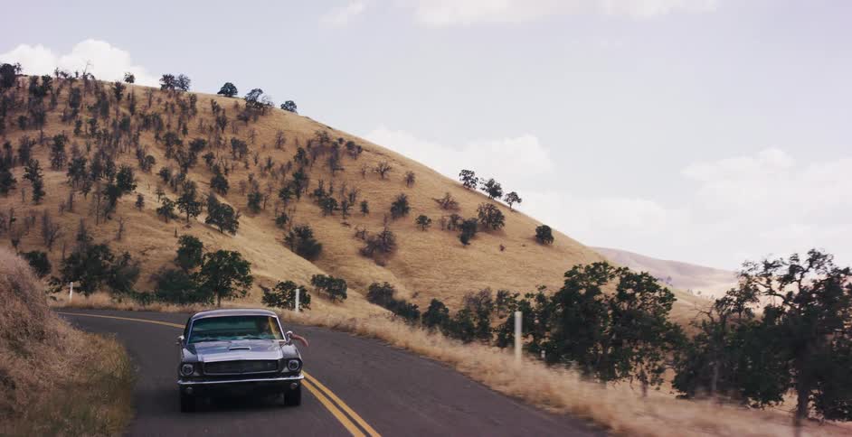 A classic grey muscle car drives along a winding road through the dry, golden hills of the countryside