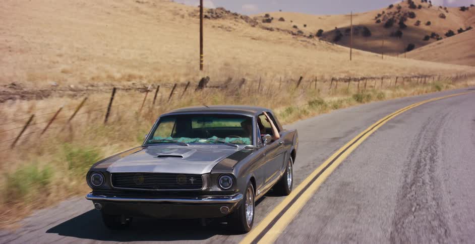 A classic silver Ford Mustang drives along a winding country road through golden, sun-drenched hills