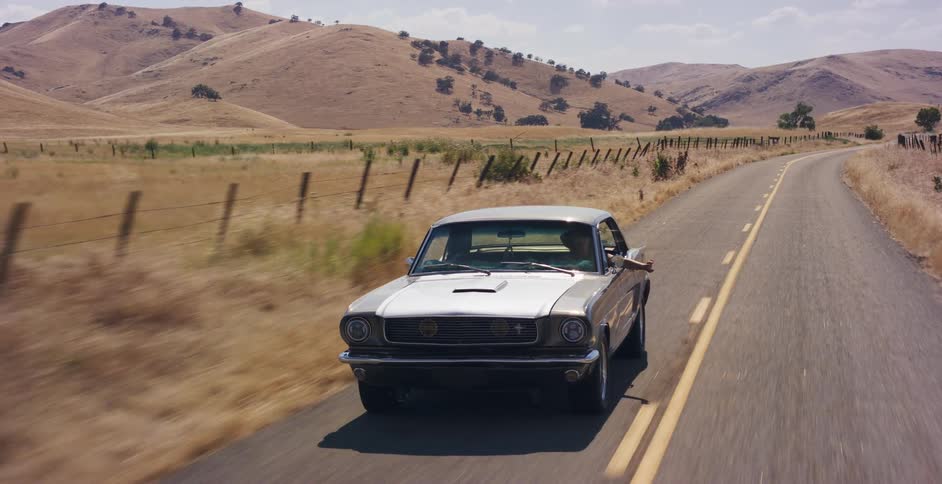 A classic silver Ford Mustang drives down a sun-drenched country road through rolling golden hills