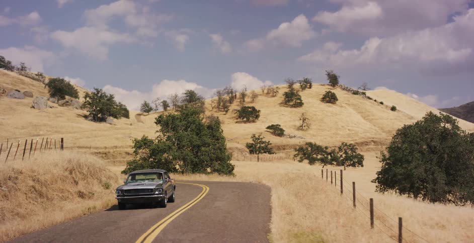 A classic gray muscle car drives along a winding road through the picturesque golden hills of the countryside