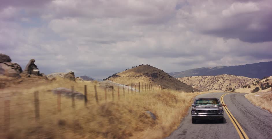 A classic silver Mustang cruises down a winding country road through golden rolling hills under a cloudy sky