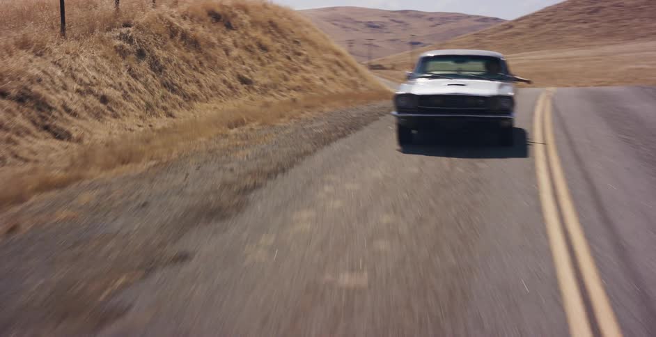 A man enjoys a scenic drive in his classic silver muscle car along a deserted road through the golden hills of the countryside