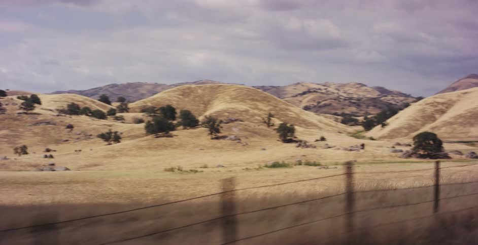 A classic muscle car drives down a lonely country road surrounded by vast, golden rolling hills