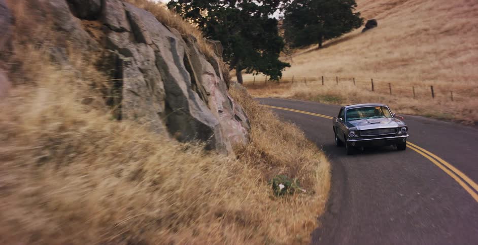 A vintage Ford Mustang takes a scenic drive along a winding country road through golden, sun-drenched hills