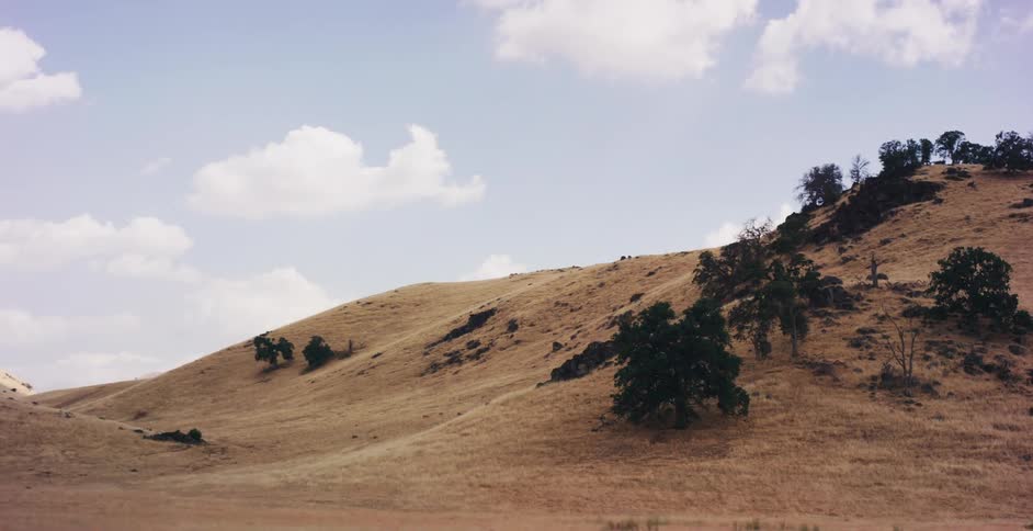 A tracking shot from a moving vehicle passes a rustic fence, revealing the vast, golden, sun-drenched rolling hills of the countryside