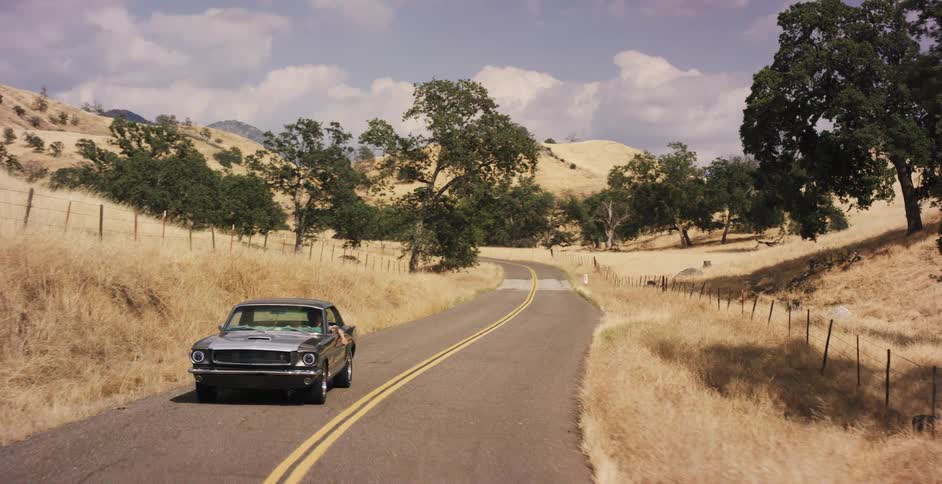 A vintage muscle car drives down a winding country road through rolling golden hills on a summer day