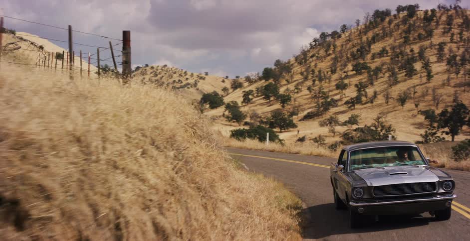A classic silver muscle car cruises along a winding country road through dry golden hills