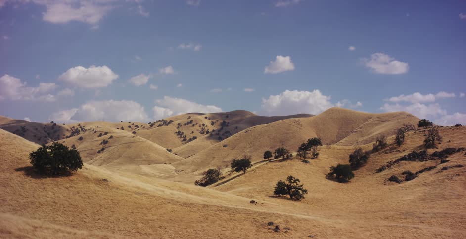 A slow pan across vast, rolling golden hills dotted with oak trees under a partly cloudy blue sky