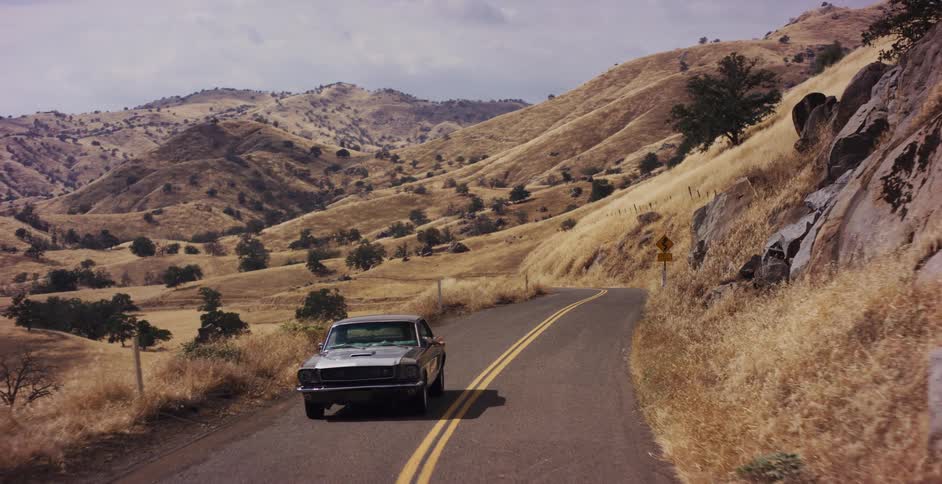Aerial view of a classic Ford Mustang driving along a winding country road through golden hills on a sunny day