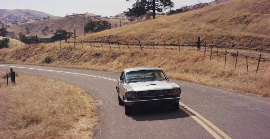 A classic silver Ford Mustang cruises in slow motion down a winding country road through dry, golden hills on a sunny day
