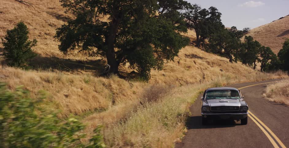A classic silver Ford Mustang takes a scenic drive along a winding country road through golden, sun-drenched hills
