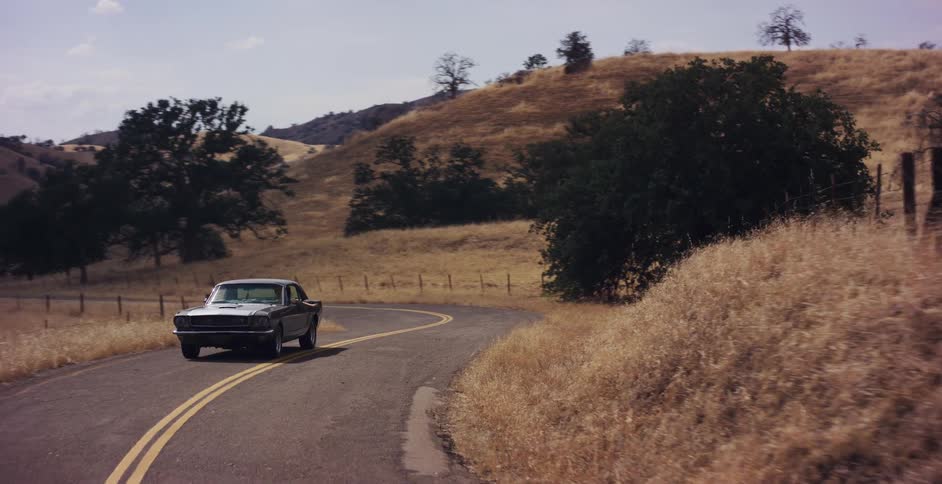 A vintage Ford Mustang cruises down a winding country road through the golden, rolling hills of California