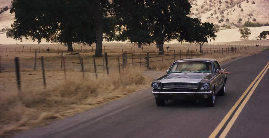 A classic dark Ford Mustang cruises along a scenic country road through rolling golden hills