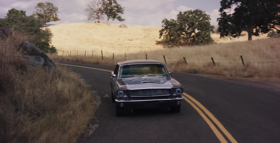 A classic Ford Mustang takes a slow, scenic drive along a winding country road through rolling golden hills