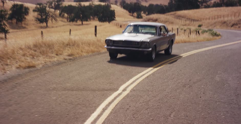 Tilting up from the speeding asphalt reveals a classic silver Mustang driving on a winding country road through sunny, golden hills