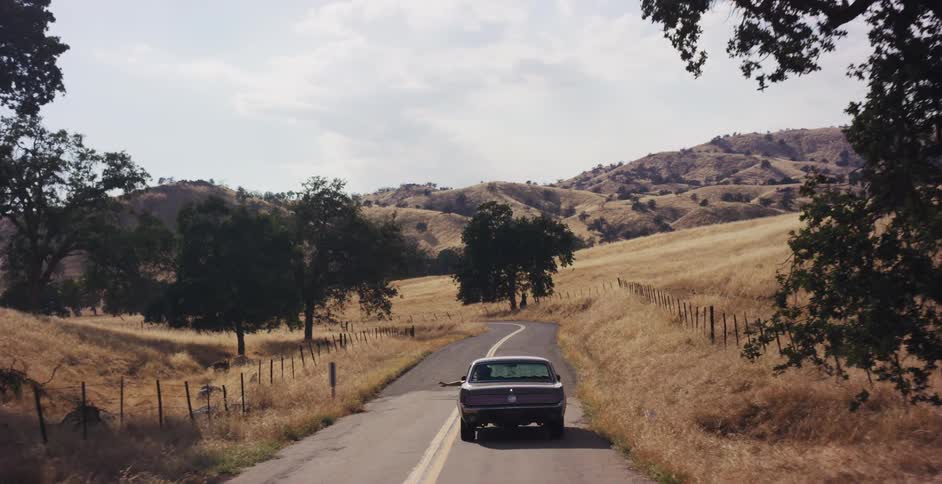 A vintage car drives down a winding country road through golden hills, with a person's hand out the window feeling the air