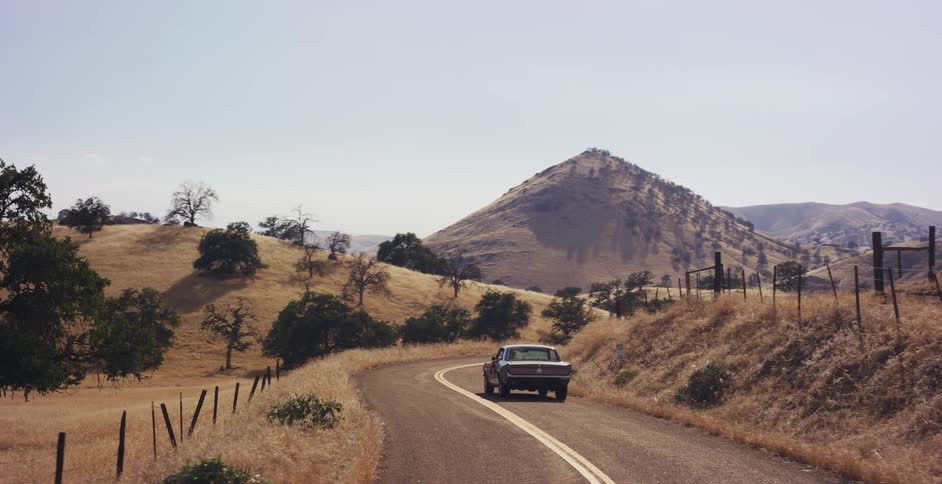 A classic American muscle car cruises down a scenic, winding country road through dry, golden rolling hills