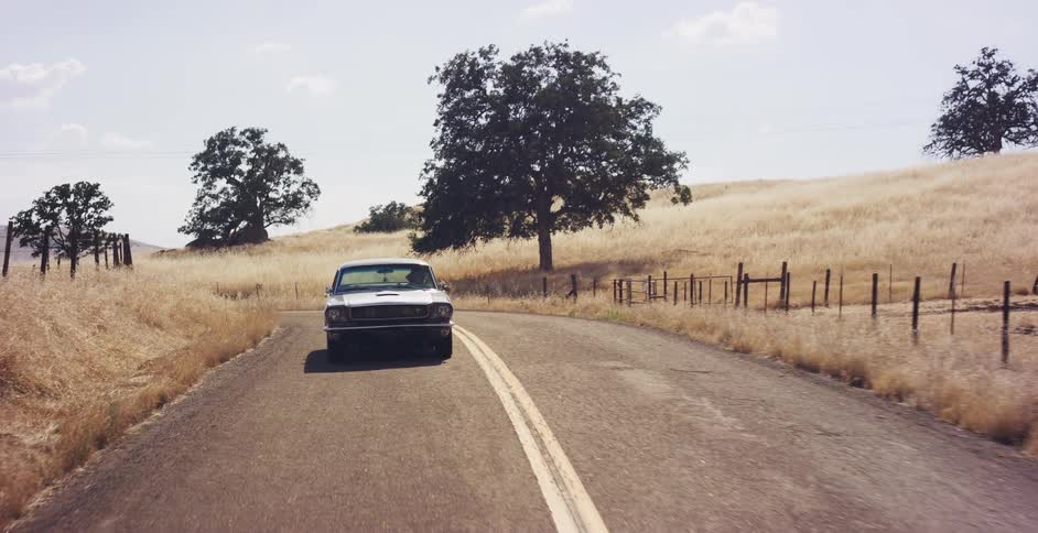 A classic silver Mustang drives along a winding country road through golden, sun-drenched hills