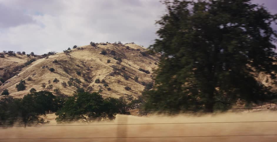A scenic drive-by view of rolling golden hills dotted with green trees under a cloudy sky in slow motion