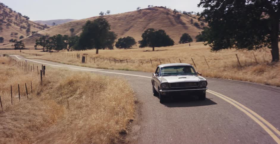 A vintage silver Ford Mustang cruises along a winding country road through sun-drenched golden hills