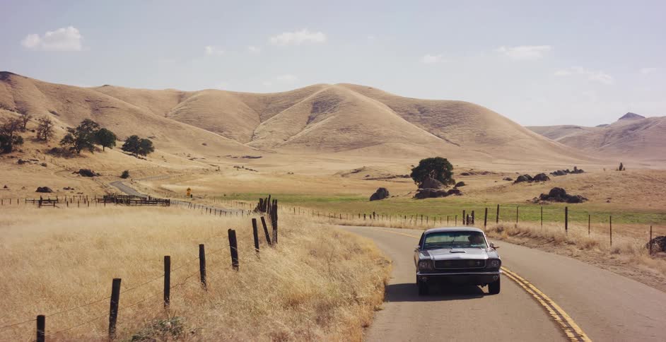 An aerial view tracks a vintage silver Ford Mustang driving along a winding country road through a vast, sun-drenched landscape of golden rolling hills
