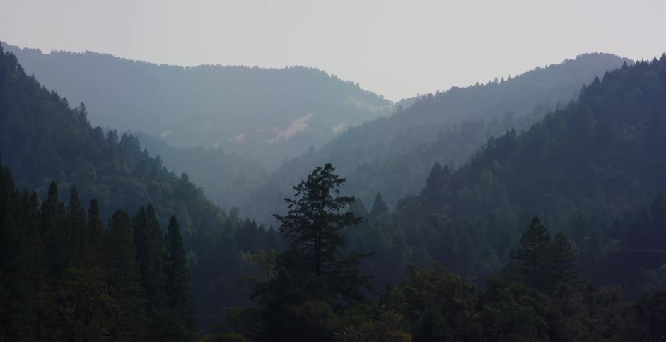 An atmospheric aerial view of a hazy, forested mountain valley reveals its layered beauty as the camera pans left