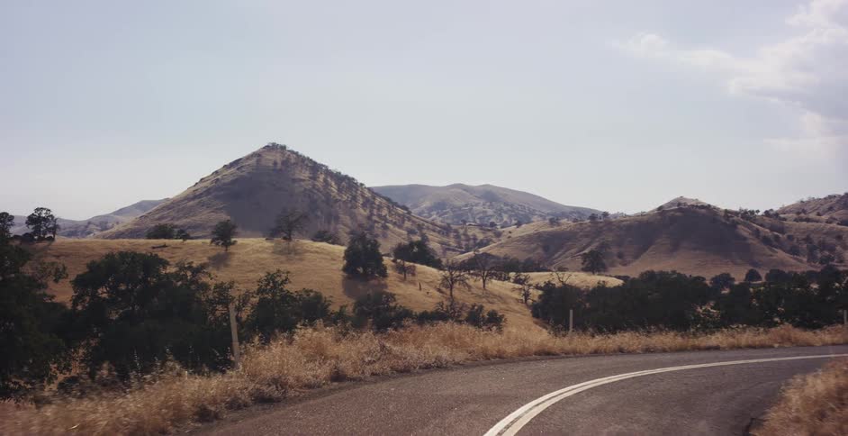 A scenic drive follows a vintage car along a winding road through the vast, golden hills of the California countryside