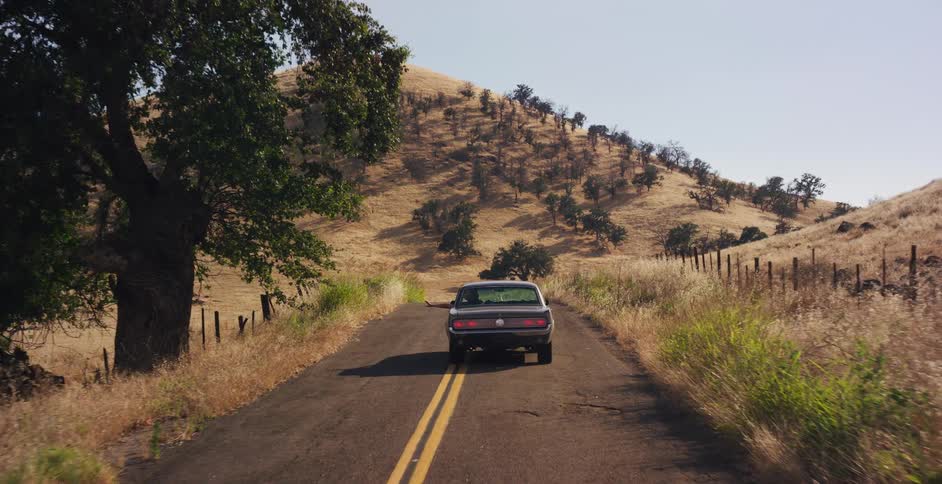A classic American muscle car cruises along a winding backroad through the golden, sun-drenched hills of the countryside