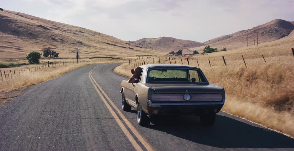 A woman in a cowboy hat enjoys a scenic drive in a classic Ford Mustang along a winding country road through golden hills