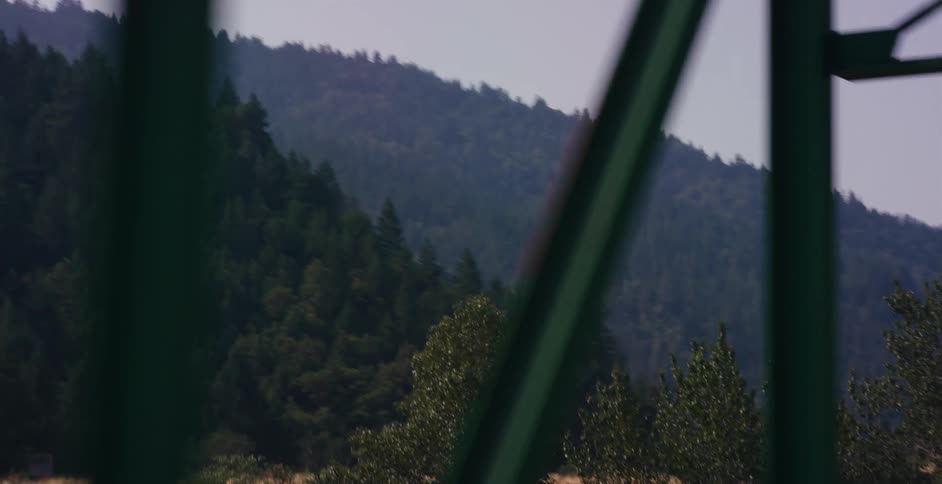 A slow-motion tracking shot from a vehicle crossing a green steel truss bridge, with a beautiful evergreen forest visible in the background