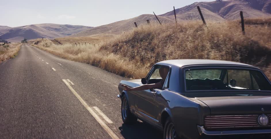 A man enjoys a leisurely drive in a classic gray muscle car, his arm resting out the open window as he cruises down a sunny country road