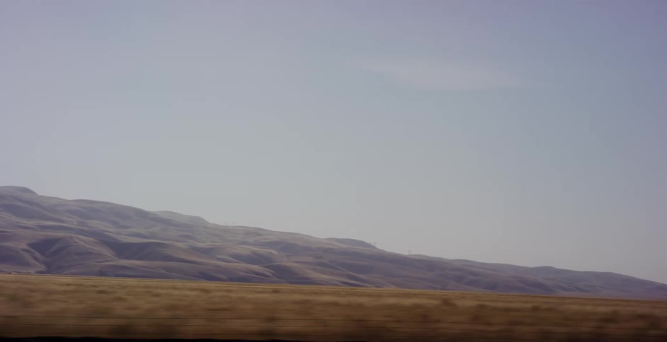 A scenic POV shot from a moving car captures the dry, golden fields and rolling hills of a hazy desert landscape as a semi-truck drives past
