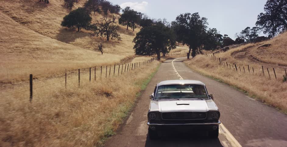 Aerial view following a classic silver Ford Mustang as it drives along a winding country road through sunny, golden hills, eventually joined by a group of motorcyclists