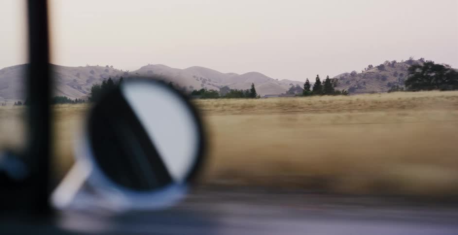The rural landscape of rolling golden hills and fields passes by the window of a moving car