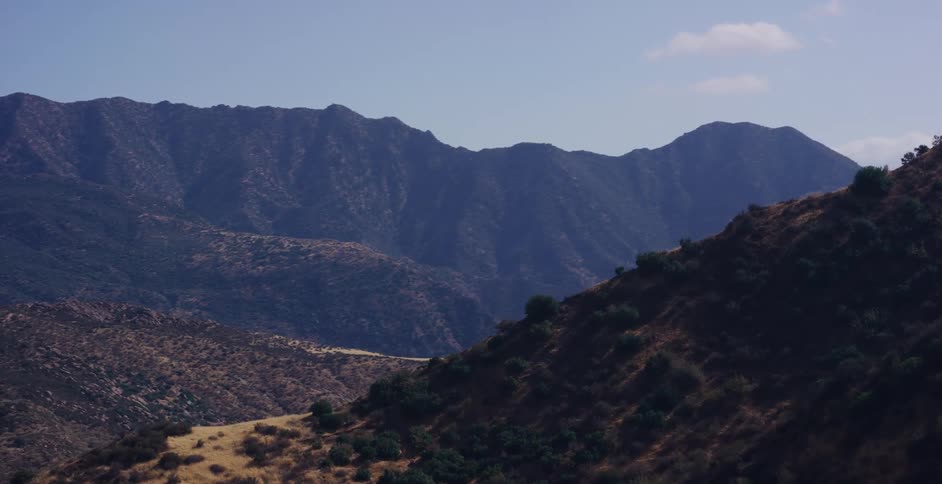 A slow zoom out reveals a vast, sunlit mountain range and dry, grassy hills on a clear day