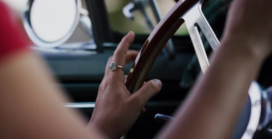 A close-up over-the-shoulder shot of a woman's hands tapping on the wooden steering wheel of a classic car