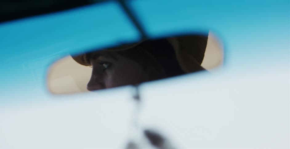A woman's eyes are seen reflected in a car's rearview mirror as she drives on a sunny day