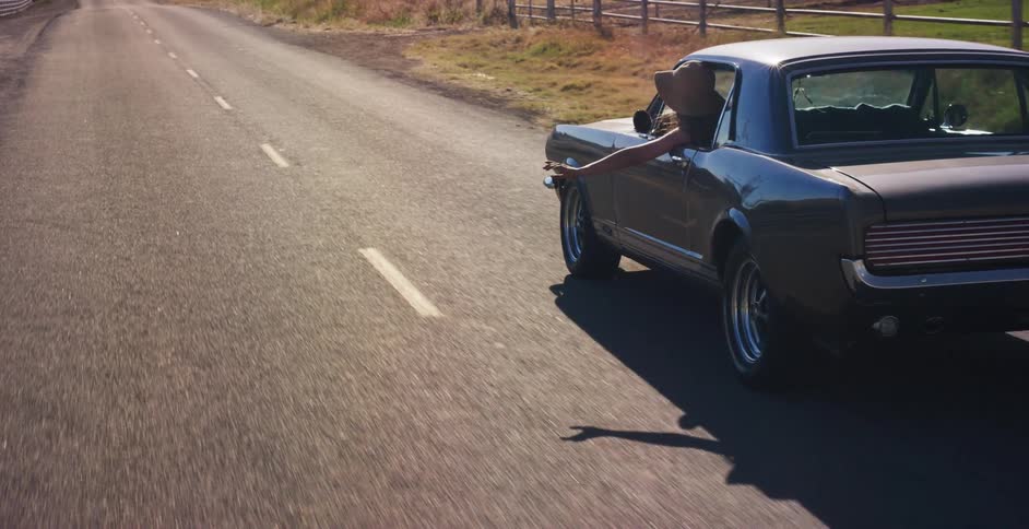 A woman in a floppy hat hangs her arm out the window of a classic muscle car while cruising down a sunny country road in slow motion