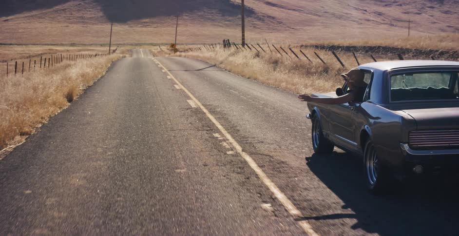 A woman with her arm out the window cruises down a sun-drenched country road in a classic American muscle car
