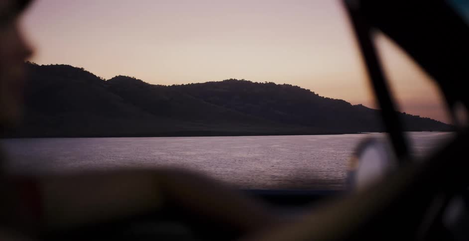 Rack focus from a woman driving a car at dusk to the tranquil lake and mountains outside her window