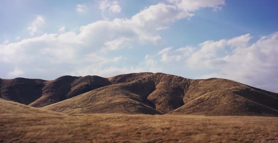 A scenic drive-by shot of vast, golden rolling hills under a beautiful blue sky with fluffy white clouds