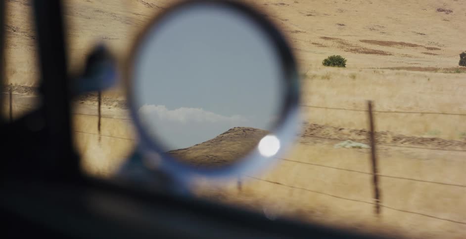 A reflection of a blue sky and golden hills is seen in the side mirror of a car driving through the countryside