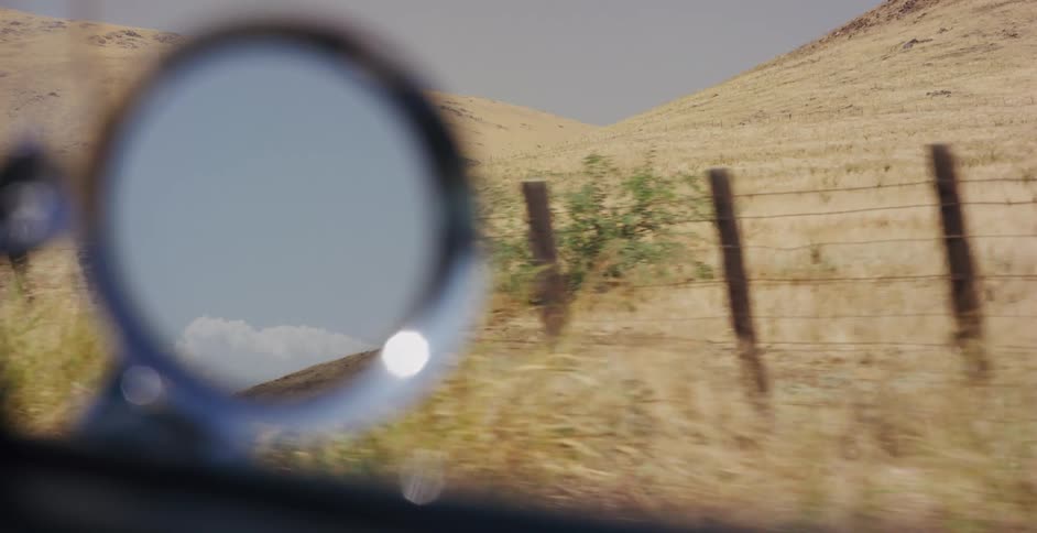 A car's side-view mirror reflects the blue sky as it drives past a dry, golden hillside with a rustic fence