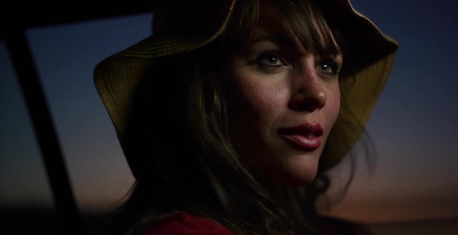 A pensive young woman wearing a sun hat sits in a car, her face illuminated by the last light of dusk