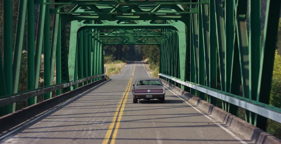 A classic muscle car is followed as it drives along a winding country road and across a distinctive green steel bridge on a sunny day